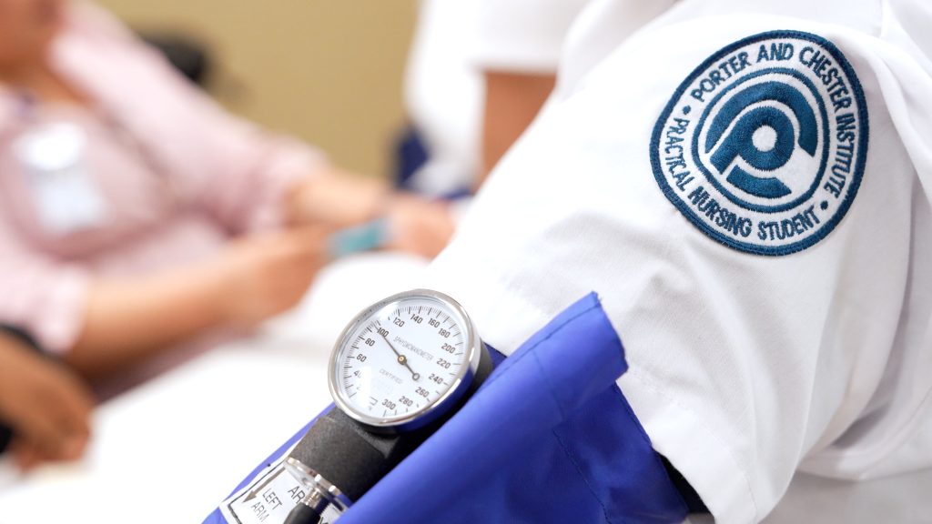 Close-up of a student wearing a Porter and Chester Institute nursing uniform with a blood pressure cuff and gauge, while another person is seated in the background.