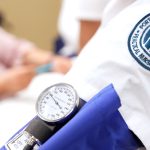Close-up of a student wearing a Porter and Chester Institute nursing uniform with a blood pressure cuff and gauge, while another person is seated in the background.