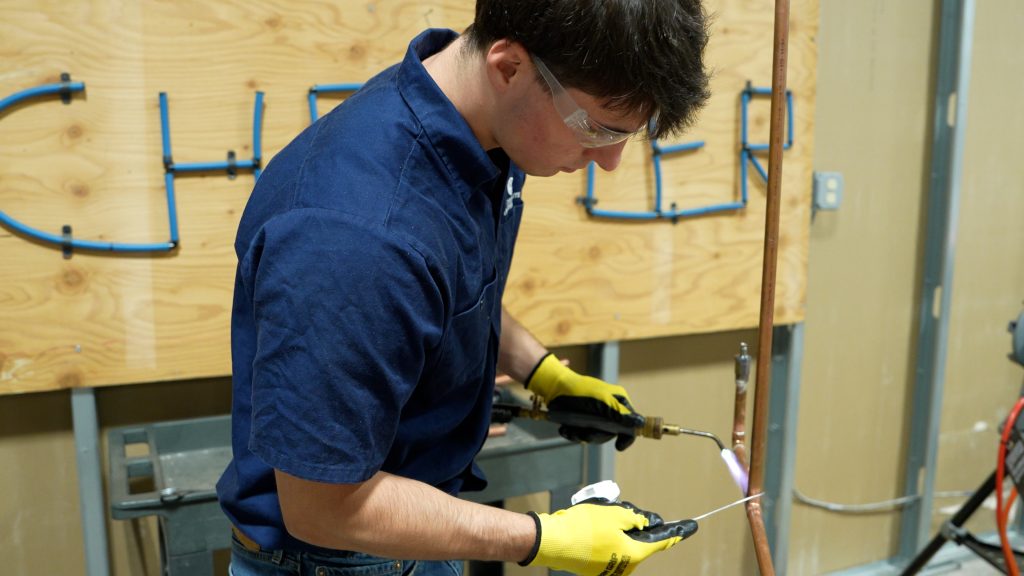 A person wearing safety glasses and gloves uses a torch to work on a copper pipe, with tools and a wooden board in the background.