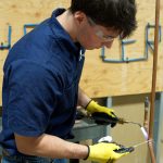 A person wearing safety glasses and gloves uses a torch to work on a copper pipe, with tools and a wooden board in the background.