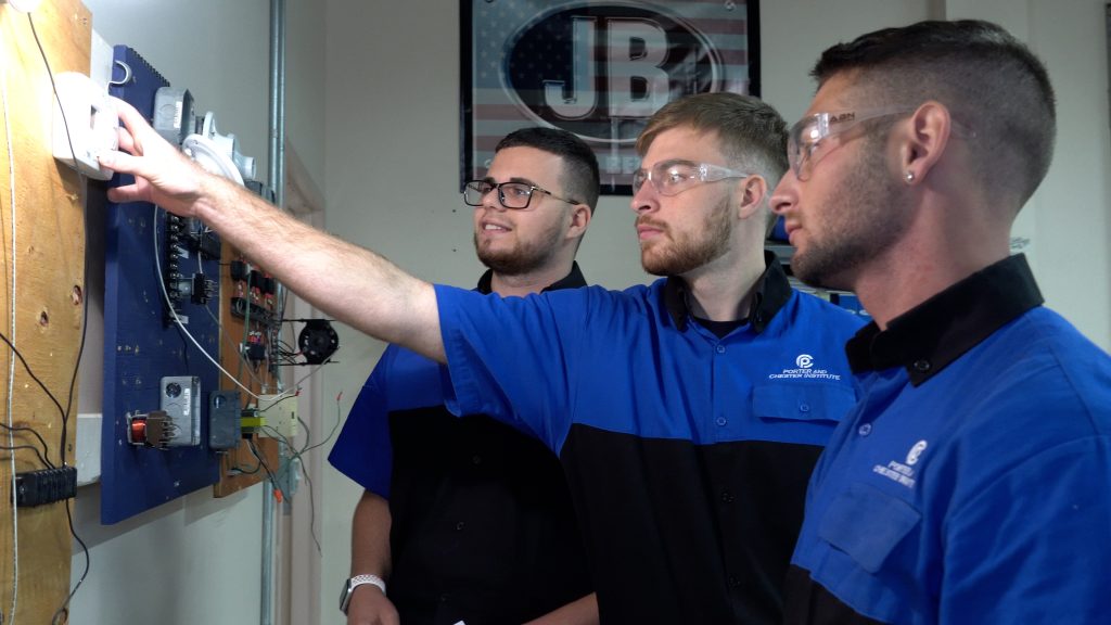 Three men wearing safety glasses and blue uniforms work on an electrical panel, focusing on a component. A sign with JB is visible in the background.