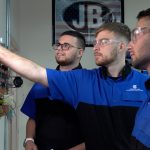 Three men wearing safety glasses and blue uniforms work on an electrical panel, focusing on a component. A sign with JB is visible in the background.