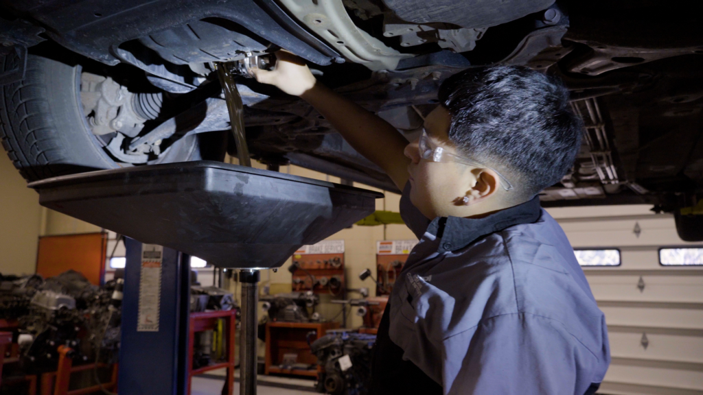 Person performing an oil change on a lifted vehicle, with a drip pan below, in a garage setting.