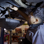 Person performing an oil change on a lifted vehicle, with a drip pan below, in a garage setting.