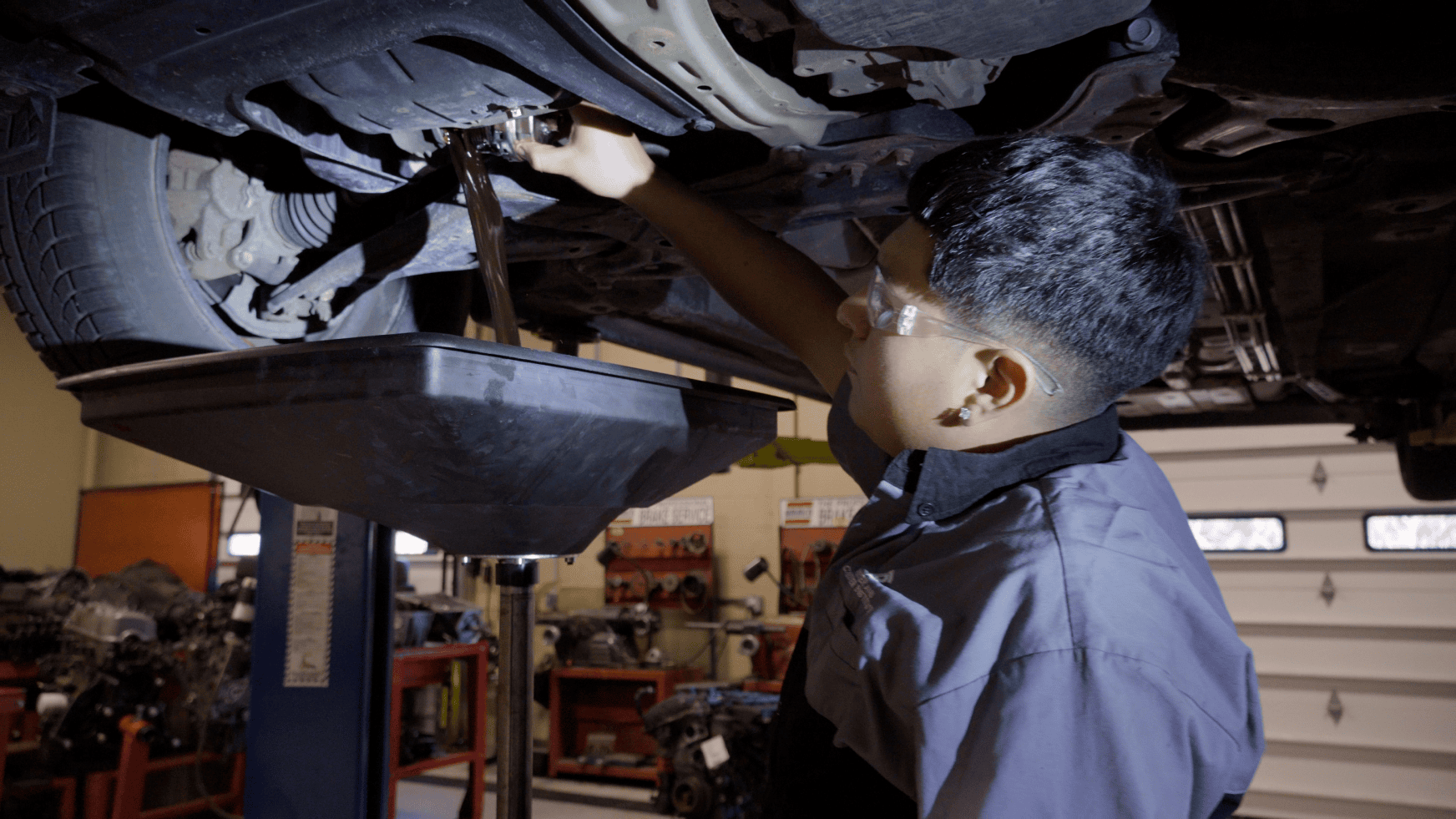 Person performing an oil change on a lifted vehicle, with a drip pan below, in a garage setting.