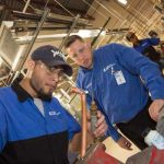 Two workers in blue shirts are focused on a piece of machinery in an industrial setting. One is handling a tool and a copper pipe, while the other observes closely. Safety glasses and protective gear are worn by both.