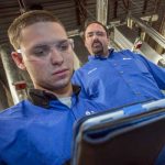 Two men in blue work shirts and safety glasses look at a tablet in an industrial setting.