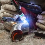 Person welding stainless steel pipes in a workshop, wearing protective gloves and using a welding torch emitting a bright light.