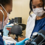 Two healthcare workers wearing masks and gloves are taking a blood pressure measurement in a clinic setting.
