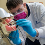 A dental student wearing protective gear practices a procedure on a dental model using instruments, focused on technique.