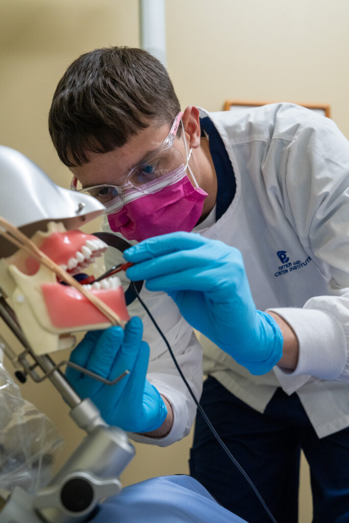 A dental student wearing protective gear practices a procedure on a dental model using instruments, focused on technique.