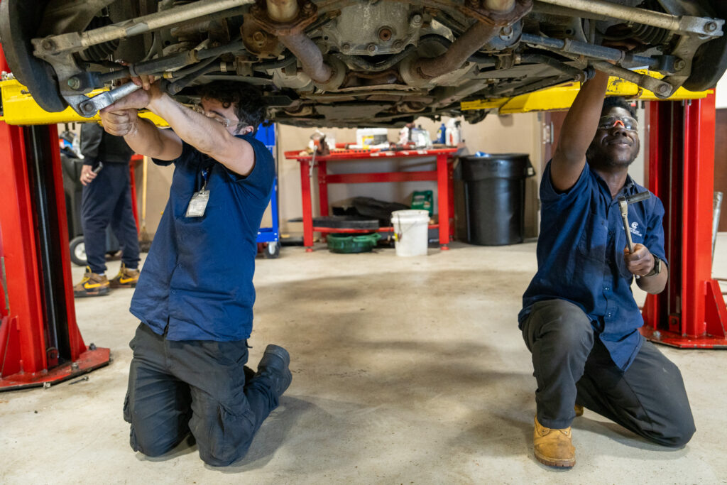 Two mechanics working under a lifted vehicle in a garage, adjusting parts. One is using a tool while kneeling, the other reaches upward. Red supports and various tools are visible in the background.