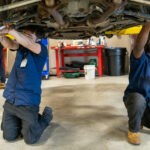 Two mechanics working under a lifted vehicle in a garage, adjusting parts. One is using a tool while kneeling, the other reaches upward. Red supports and various tools are visible in the background.