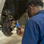 Mechanic in a blue shirt using a wrench to work on a cars brake system. Safety glasses and pen tucked in his ear. Car raised on a lift in a workshop.
