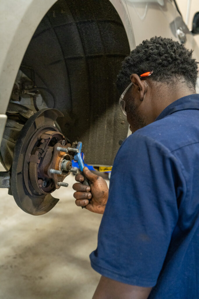 Mechanic in a blue shirt using a wrench to work on a cars brake system. Safety glasses and pen tucked in his ear. Car raised on a lift in a workshop.